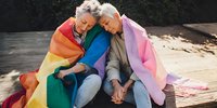 Senior Asian couple sitting close together outdoors, wrapped in rainbow and pink pride flags, holding hands and sharing intimate moment of connection and acceptance.
