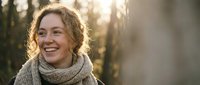 Happy young woman with curly hair wearing knit scarf laughing outdoors in golden sunlight. Natural portrait for lifestyle and wellness content.