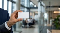 Businessman in dark suit holding blank white business card in modern office environment with blurred background for corporate branding.