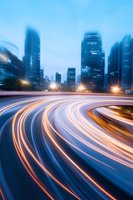 Modern cityscape with light trails from traffic against illuminated skyscrapers at dusk, creating dynamic curves and motion blur effect in blue tones.
