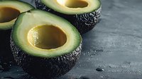 Fresh halved avocados with pits removed, displaying creamy green flesh on dark textured surface. Suitable for food, nutrition, healthy eating, recipe, and culinary design projects.