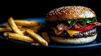 Juicy cheeseburger with bacon, lettuce, tomato and melted cheese on sesame seed bun served with golden french fries on dark plate, low key food photography.