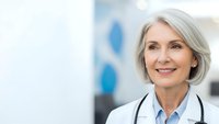 Confident mature female doctor with silver hair and stethoscope smiling in modern medical office setting for healthcare and wellness campaigns.