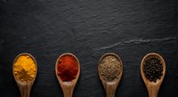 Four wooden spoons with colorful spices on dark slate background, turmeric, paprika, cumin, black pepper, top view, copy space.