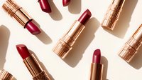 Luxurious rose gold lipsticks in various shades of red and burgundy scattered on white background with dramatic shadows, creating modern minimalist makeup composition.