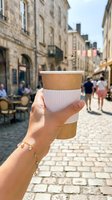 Hand holding takeaway coffee cup in European street with historic stone buildings and pedestrians in background, ideal for beverage, travel, and lifestyle marketing.