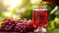 Red grape juice pouring into glass with fresh grapes on wooden table in sunlight for healthy beverage and natural drink concepts.