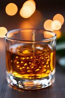 Pouring golden liquid into glass tumbler with bubbles and bokeh lights in background, close-up shot showing rich amber color and crystal clarity.