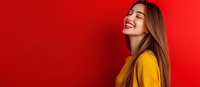 Young Caucasian woman with long brown hair wearing yellow sweater laughing with closed eyes against bright red background, expressing joy and happiness.