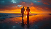 Romantic couple holding hands walking on beach at sunset with dramatic orange sky and ocean waves reflecting golden light on wet sand.