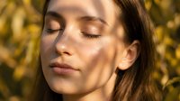 Young woman with fair skin and long brown hair, eyes closed in peaceful expression, golden hour sunlight casting warm glow on face, outdoor natural lighting for wellness and beauty content.