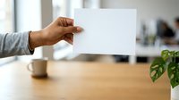 Male hand holding blank white card in modern office workspace with wooden desk, coffee cup, and green plant in background.