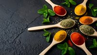 Colorful spices and herbs in wooden spoons arranged on dark slate background. Fresh mint leaves accent vibrant turmeric, paprika and dried seasonings.