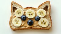 Creative breakfast toast shaped as cat face with peanut butter, banana slices and blueberries on white background. Fun food art for kids.