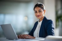 Professional South Asian businesswoman working on laptop in modern office environment smiling confidently at camera wearing navy blazer.