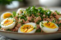 Fresh tuna salad with soft-boiled eggs cut in halves, garnished with chopped parsley and black pepper on white plate, close up view of healthy meal.