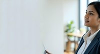Young South Asian woman in professional business attire holding tablet, working in modern office environment with natural light and plants, suitable for corporate, technology, and business concepts.