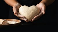 Baker hands holding heart-shaped bread dough with flour dust in warm kitchen lighting for homemade baking and culinary love concept.
