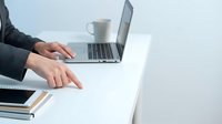Professional businesswoman typing on laptop computer at modern white desk workspace with coffee cup and notebooks for remote work productivity.