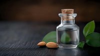 Glass bottle with cork stopper containing clear liquid, almonds and green leaves on dark wooden surface for natural skincare and aromatherapy products.