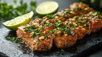 Grilled salmon fillet pieces glazed with teriyaki sauce, garnished with fresh herbs, chopped green onions and lime wedges served on black stone plate.