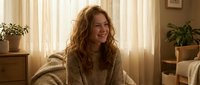 Young Caucasian woman with curly red hair smiling warmly in cozy knit sweater sitting in sunlit living room with plants and soft natural lighting for lifestyle content.