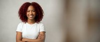 Young African American woman with curly red hair smiling confidently with arms crossed wearing white t-shirt in modern office environment for business content.