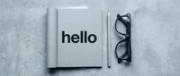 Open notebook with bold hello text beside pencil and glasses on concrete surface, minimalist monochrome workspace flat lay concept.