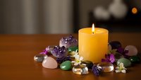 Burning yellow candle surrounded by colorful gemstones, crystals and purple flowers on wooden surface creating peaceful meditation atmosphere.