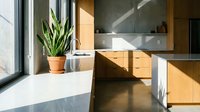 Modern kitchen with wooden cabinetry, white countertops, and natural light streaming through large windows, featuring potted plant and minimalist design for contemporary home interiors.
