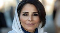 Middle Eastern woman wearing white hijab smiles warmly at camera with confident expression, natural makeup, and dark hair visible, against soft blurred background.