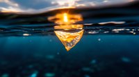 Split view underwater photo of glowing paper boat floating at sunset, with dramatic sky reflection on dark blue ocean surface creating magical atmosphere.