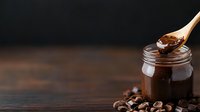 Rich chocolate spread in glass jar with wooden spoon and cocoa beans on rustic wooden table against dark background for food photography and culinary designs.