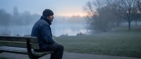 Thoughtful middle-aged man sitting alone on park bench during misty dawn, contemplating by lake. Peaceful morning solitude and reflection concept.