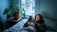 Young woman relaxing in cozy bedroom using smartphone near window with houseplants on sunny morning for social media and communication lifestyle.