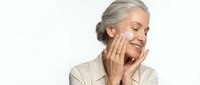 Mature woman with gray hair touching face gently, skincare routine concept on white background. Anti-aging beauty treatment and wellness lifestyle.