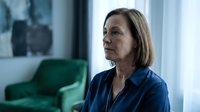 Thoughtful senior Caucasian woman in blue blouse sitting indoors, looking away with serious expression in modern living room.