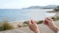 Senior woman holding blank white card at coastal beach location with sea and sandy shore in background, suitable for travel, vacation, retirement, and lifestyle marketing campaigns.