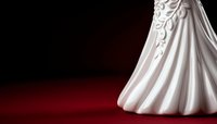 White wedding dress with elegant floral lace detail and flowing train on dark red velvet background, dramatic studio lighting creates romantic bridal atmosphere.