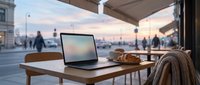 Laptop on wooden desk by window overlooking urban street at sunset, with croissant and sweater, workspace for remote work or freelance projects.