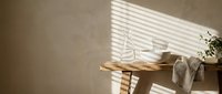 Young woman with light skin tone, wearing casual clothing, sitting at wooden table in minimalist interior with natural sunlight and window blinds creating striped shadows, suitable for lifestyle and wellness content.