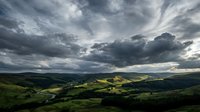 Dramatic stormy sky with rolling green hills and valleys below, moody clouds casting shadows across lush countryside landscape for nature and weather backgrounds.