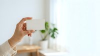 Hand holding blank white business card in bright interior space with blurred green plant and natural window light, suitable for branding and corporate identity mockups.