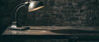Vintage desk lamp with curved metal arm casting warm light on wooden surface against aged brick wall backdrop.