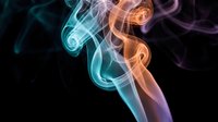 Colorful smoke swirls in teal and orange tones against a dark black background, creating an abstract, artistic flow of light and motion.