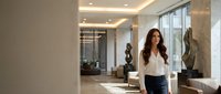 Professional businesswoman with long dark hair standing in modern corporate office lobby with marble columns and contemporary lighting for workplace concepts.