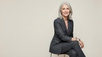 Confident mature businesswoman with silver hair in dark suit sitting professionally against neutral background for corporate and leadership concepts.