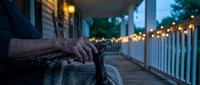 Elderly hands resting on wicker basket during peaceful evening on illuminated porch with warm string lights creating cozy outdoor atmosphere.
