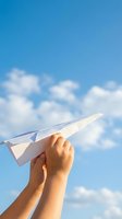 Hand holding white paper airplane against bright blue sky with fluffy white clouds symbolizing dreams, freedom, childhood memories and aspirations.