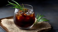 Cold brew coffee cocktail garnished with fresh rosemary sprigs and ice cubes served in round glass on rustic burlap napkin against dark background.